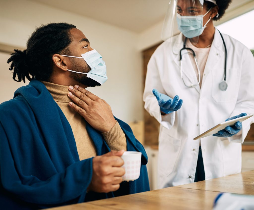African American man wrapped in blanked talking to his doctor and complaining about sore throat during a home visit. Both of them are wearing face masks due to coronavirus pandemic.